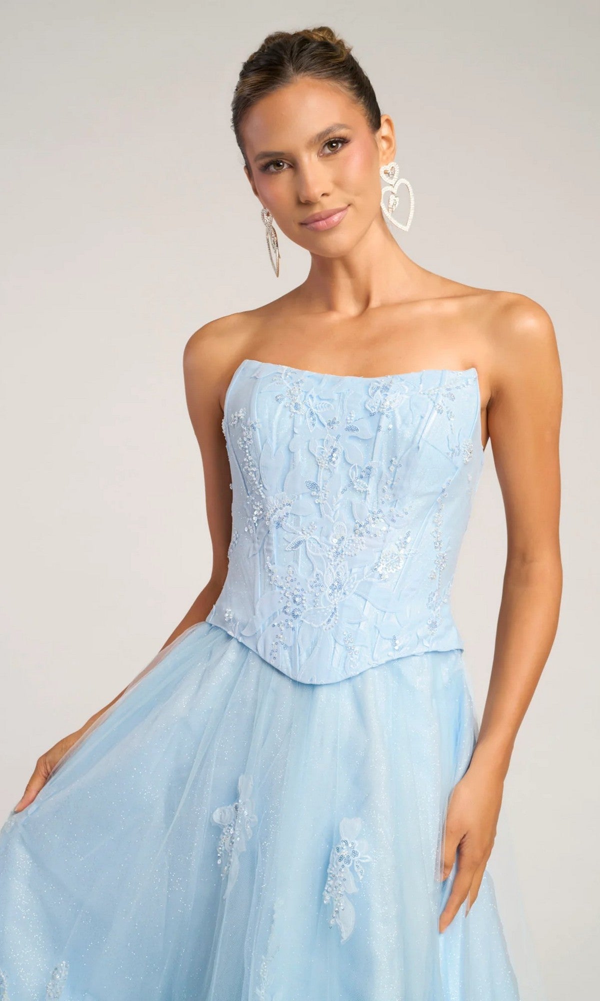 A woman in the Portia and Scarlett PS26061 long formal dress, featuring a strapless, light blue embroidered A-line design with a fitted corset bodice and flared skirt, stands against a plain background with her hair tied back and large heart-shaped earrings.